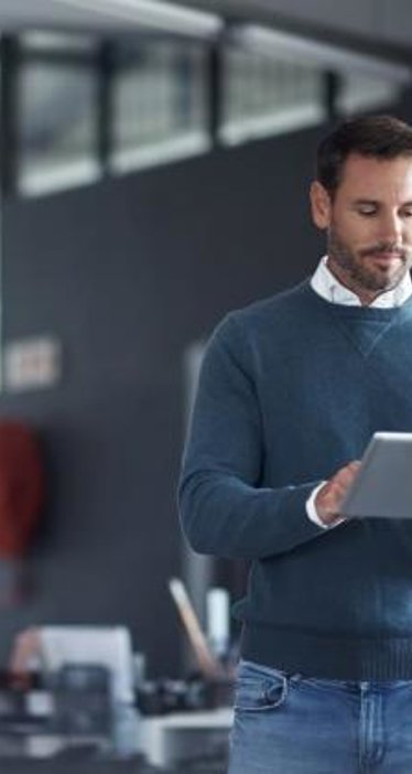 Person in blue sweater and jeans using a tablet in an office with desks and chairs; face pixelated for privacy.